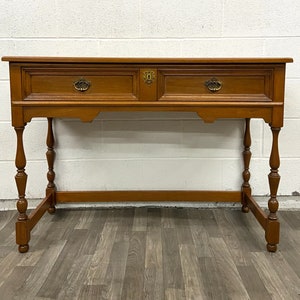 May include: A wooden console table with two drawers and turned legs. The table has a light brown finish and brass hardware.