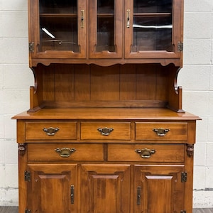 May include: A medium-brown wooden hutch featuring an upper cabinet with three glass-paneled doors and interior shelving. The base includes three top drawers, two larger lower drawers, and three lower cabinet doors, all accented with brass hardware.