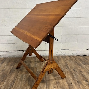 May include: A vintage wooden drafting table with an adjustable angled top. The table is constructed from light brown wood and features a sturdy, angled base. The tabletop is approximately 36 inches wide and 24 inches deep.