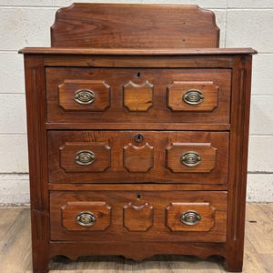 May include: A three-drawer wooden chest with a dark brown finish. The chest features brass hardware and decorative panels on each drawer. The top of the chest has a raised back. The chest is a classic piece of furniture.