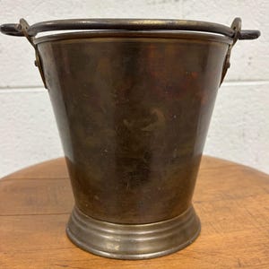 May include: A vintage metal bucket with a bronze finish and a sturdy handle. The bucket has a flared shape and a decorative base. The handle is made of a darker metal. The bucket is likely used for decorative purposes.