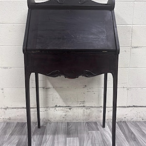 May include: A dark brown wooden secretary desk featuring a slanted drop-front lid, a single drawer below the writing surface, and four slender legs. The top gallery is decorated with carved leaf motifs, and the front panel has a keyhole.