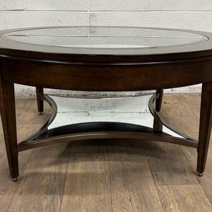 May include: A dark brown oval coffee table with a clear glass top. The table has a wooden frame and legs, with a metal support structure. The table is approximately 48 inches long and 24 inches wide.