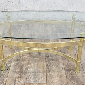 May include: Oval glass top coffee table with a gold metal frame. The frame has a unique design with curved legs and a crossbar.