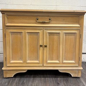 May include: A light-colored wooden cabinet with a drawer and two doors. The cabinet has a rectangular shape with a decorative top and base. The drawer has a brass handle, and the doors have brass knobs. The wood grain is visible.