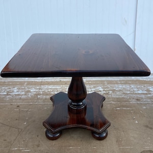 May include: A dark brown square wooden table with a pedestal base. The table has a smooth, polished finish and a simple, elegant design.