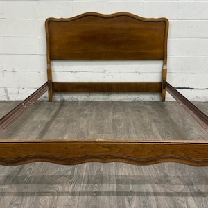May include: A wooden bed frame featuring a tall headboard with a curved, scalloped top edge and a matching low-profile footboard. The frame includes side rails and a warm, medium-brown wood grain finish with smooth, flowing lines throughout.