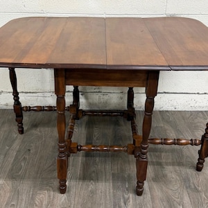 May include: A brown wooden drop-leaf table featuring a rectangular top with rounded corners and two hinged side leaves. The table is supported by six turned legs connected by horizontal stretchers, providing a stable base for the surface.
