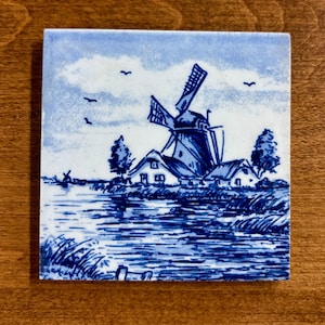 May include: A square ceramic tile featuring a blue and white hand-painted scene of a traditional Dutch windmill situated beside a calm river. The landscape includes small houses, trees, and birds flying in the sky, rendered in classic delft blue.