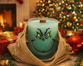 May include: A frosted glass candle in a light green hue, featuring a black graphic of a mischievous face with yellow eyes and a wide grin. The candle has a wooden wick and is wrapped in a textured tan burlap fabric, creating a rustic appearance.