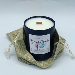 May include: Black candle with a white wax top and a wooden wick. The candle is in a burlap bag and has the text "Lit Wicks London Baby Gender Reveal Hand Poured Soy Wax @litwickslondonstore" on the side.