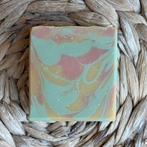 May include: A rectangular bar of handmade soap with a swirled design. The soap features a blend of colors, including light green, pink, and yellow. The soap has a textured appearance, suggesting natural ingredients.