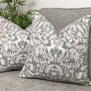 May include: Two grey throw cushions featuring a white botanical and deer design. The square cushions rest on a grey sofa. The pattern includes leaves, flowers, and deer.