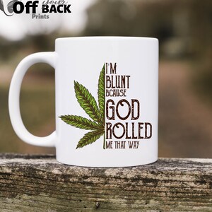 May include: White ceramic mug featuring a green cannabis leaf and the brown text "I'M BLUNT BECAUSE GOD ROLLED ME THAT WAY". The mug has a curved handle and the words "Off Your Back Prints" are at the top left.