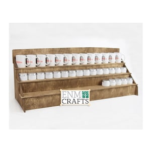 May include: A wooden shelf unit with four tiers, designed to display mugs and small jars. The shelf is stained a dark brown color and has a rustic appearance. The top tier holds a row of white mugs with red lettering. The lower tiers hold rows of small silver jars.