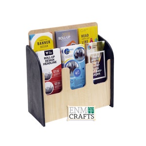 Peut inclure: Un porte-brochures en bois avec quatre fentes pour afficher des brochures, des dépliants ou des brochures. Le porte-brochures a une base noire et un dessus en bois naturel. Le texte sur les brochures comprend "Roll-Up Design Headline", "2018 Roll-Up Template", "Sample Text Banner Design", et "Head Sample Text Line". Le porte-brochures porte le texte "ENM CRAFTS" imprimé sur le devant.