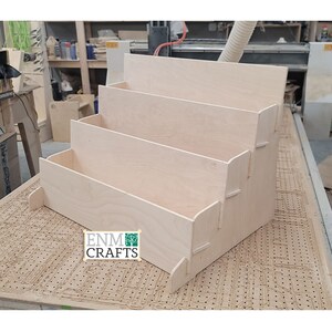 Puede incluir: Un expositor de madera de tres niveles. El expositor está hecho de madera clara y tiene un acabado natural. El expositor está etiquetado con "ENM CRAFTS".