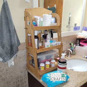 May include: A wooden bathroom shelf with three tiers, holding various toiletries and personal care items, including bottles, jars, and containers. The shelf is in a bathroom setting with a white sink and a gray tiled wall.