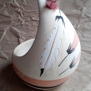 May include: Cream-colored ceramic vessel with a curved shape, featuring a white textured feather design and pink accents. The vessel has a small opening and a decorative pink tassel at the top. Black lines and a pink band adorn the exterior.