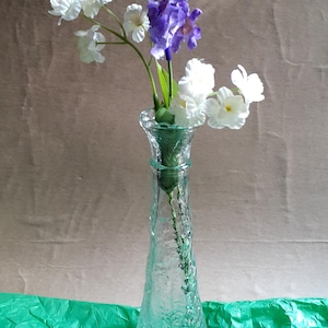 May include: A clear glass vase with a textured surface holds a bouquet of white and purple flowers. The vase has a flared shape and sits on a green surface. The background is a neutral brown.