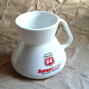 May include: White ceramic pitcher with a bulbous base and a handle. The pitcher features the Phillips 66 logo in red and white, with the words "SuperClean Unleaded Gasolines" below.