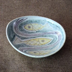 May include: A shallow, oval-shaped ceramic bowl with a light blue and green mottled glaze. The interior features three hand-painted yellow fish with cross-hatched scales, arranged in a flowing, wavy pattern defined by dark purple lines.