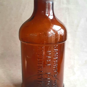 May include: A vintage amber glass bottle with embossed text. The bottle has a rounded shoulder and a narrow neck. The text reads "Sanford's Ink, Half Pint, And Library Paste". The bottle is a warm brown color.