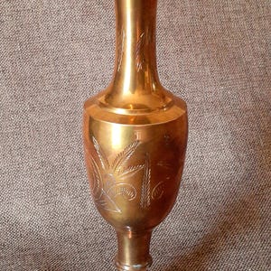 May include: A tall, gold-colored brass vase with an etched design of leaves and stems. The vase has a wide base and a narrow neck.