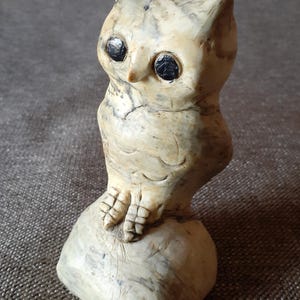 May include: A white ceramic owl figurine with black eyes, perched on a small, round base. The owl has a detailed, textured surface and a slightly tilted head.