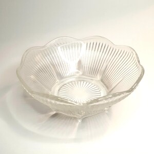 May include: A clear glass serving bowl with a scalloped edge and vertical ribbed detailing. The bowl has a starburst pattern at the base and is designed with a flower-like shape. The bowl is ideal for serving salads, fruits, or snacks.