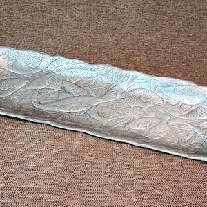May include: A rectangular silver-toned metal tray featuring an embossed design of multiple fish swimming in various directions. The surface has a textured, metallic finish with raised details depicting scales, fins, and aquatic patterns across the entire base.