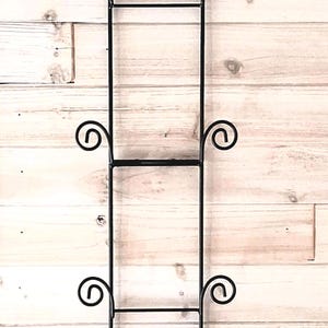 May include: A tall, vertical black metal wall rack featuring a series of rectangular frames connected by straight bars. The structure includes decorative scrollwork accents on both sides at four distinct levels for display purposes.