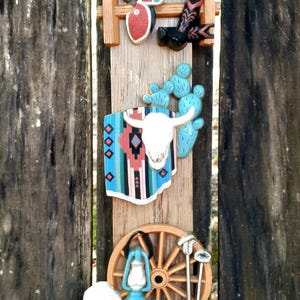 May include: Wooden wall decor featuring a cowboy theme. Includes a saddle, boots, a steer skull, a wagon wheel, a lantern, a hat, and a rope. The color palette includes red, blue, white, and brown.