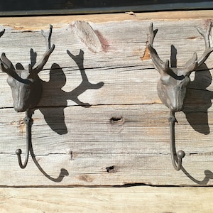 May include: Rustic wall-mounted coat rack featuring two deer head hooks. The hooks are made of dark metal and are mounted on a weathered wooden plank. The wood has a natural, aged appearance, with visible grain and knots.