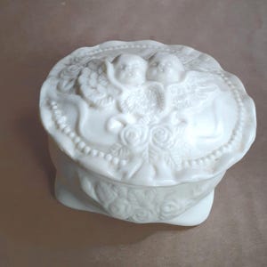 May include: A white ceramic trinket box with a lid featuring two cherubic figures with wings. The box has a beaded edge and rose details. The overall design is ornate and decorative.