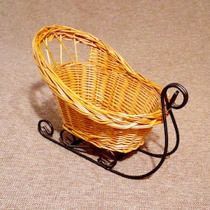 May include: A small, decorative wicker sleigh with a woven basket body and curved black metal runners. The sleigh has a rounded back and is designed for display or decorative purposes. The wicker is a light brown color.