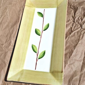 May include: Rectangular serving platter with a light green border and a white center. A painted brown stem with green leaves decorates the center. The platter is on a brown, textured surface.