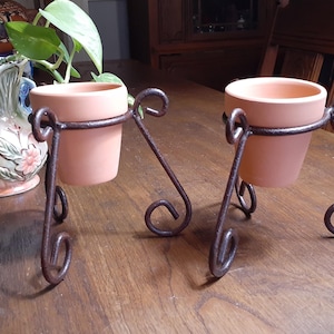 May include: Two small, terracotta-colored flower pots sit in decorative, wrought-iron stands. The stands have a dark, aged finish and feature scrolled details. A decorative vase with a floral design is visible in the background.