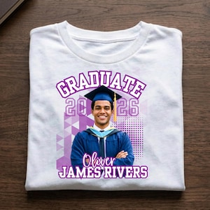 May include: White t-shirt with a graduation design. The design includes the word "GRADUATE" in purple, the year "2026", and a photo of a graduate in a cap and gown. The graduate's name, "Oliver JAMES RIVERS", is also printed on the shirt.