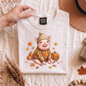 Peut inclure: Un t-shirt blanc avec une étiquette Comfort Colors présentant un dessin de cochon portant un bonnet en tricot beige et un pull à motifs patchwork. Le cochon est entouré de citrouilles orange et de feuilles d'automne sur un tissu clair.