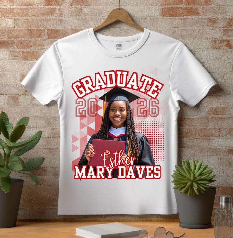 May include: White t-shirt featuring a graduation design. The design includes the word "GRADUATE" in red, the year "2026", and a photo of a smiling graduate holding a diploma. The graduate's name, "Esther MARY DAVES", is also printed on the shirt.