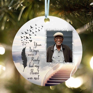 May include: A white round bauble with a photo of an older man wearing a black jacket and a tan hat. The bauble features a black feather and a staircase ascending to a cloudy sky. The text on the bauble reads "Your Wings were Ready but my Heart was not."