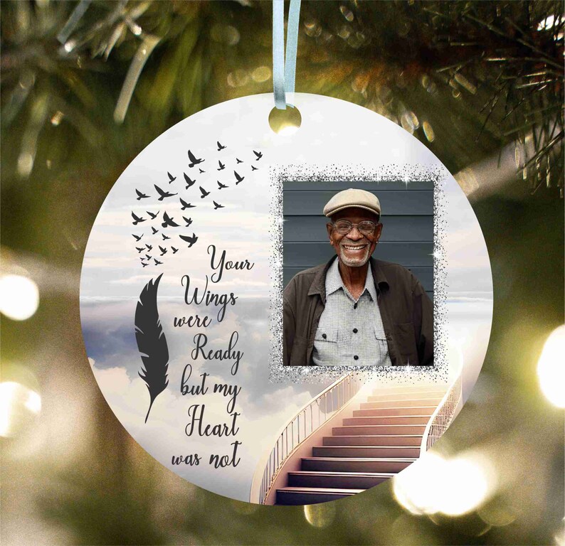 May include: A white round bauble with a photo of an older man wearing a black jacket and a tan hat. The bauble features a black feather and a staircase ascending to a cloudy sky. The text on the bauble reads "Your Wings were Ready but my Heart was not."