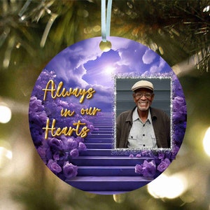 May include: A purple and white Christmas bauble featuring a photo frame with the words "Always in our Hearts". The photo frame displays a picture of an older man with a white beard and a light blue hat. The bauble is adorned with purple flowers and a stairway ascending towards a bright light.