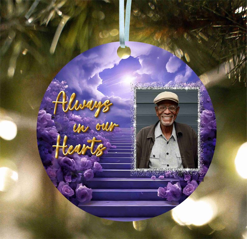 May include: A purple and white Christmas bauble featuring a photo frame with the words "Always in our Hearts". The photo frame displays a picture of an older man with a white beard and a light blue hat. The bauble is adorned with purple flowers and a stairway ascending towards a bright light.
