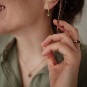 Puede incluir: Primer plano de la oreja de una persona con un pendiente de aro dorado con una peque&ntilde;a piedra verde. La persona lleva una camisa verde y un collar de oro con un peque&ntilde;o colgante de piedra verde.