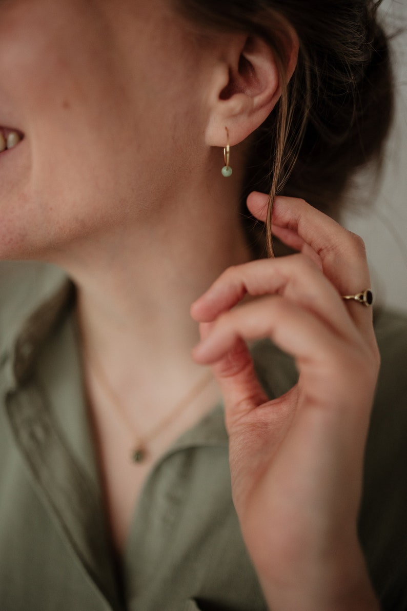 Puede incluir: Primer plano de la oreja de una persona con un pendiente de aro dorado con una peque&ntilde;a piedra verde. La persona lleva una camisa verde y un collar de oro con un peque&ntilde;o colgante de piedra verde.