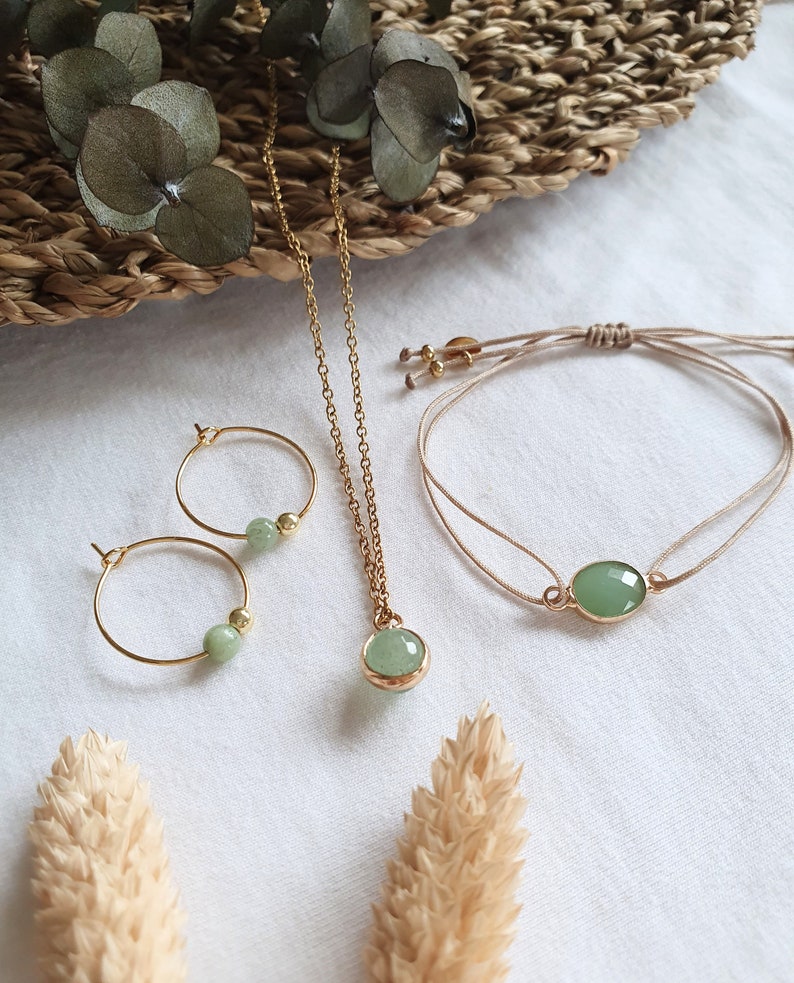 Puede incluir: Un conjunto de joyas de piedra verde que incluye un collar, una pulsera y pendientes. El collar y los pendientes tienen una cadena y aros dorados con cuentas de piedra verde. La pulsera est&aacute; hecha de un cord&oacute;n beige con un colgante de piedra verde.