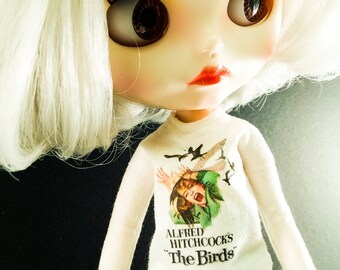 May include: A white doll with blonde hair wearing a white long-sleeved shirt with a graphic of Alfred Hitchcock's "The Birds" movie poster. The doll is wearing black and white striped pants.