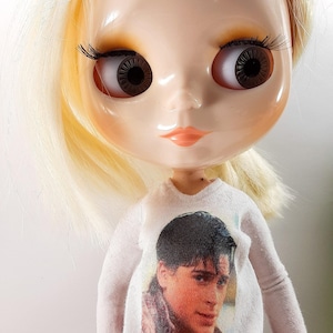 May include: A doll with blonde hair, large brown eyes, and long eyelashes. The doll is wearing a white long-sleeved shirt with a printed image of a man. The doll's face is pale with a slight blush on the cheeks and lips.